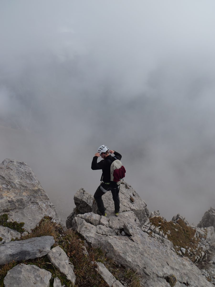 Kletterer mit Helm steht auf einem Felsgrat über einem Wolkenmeer