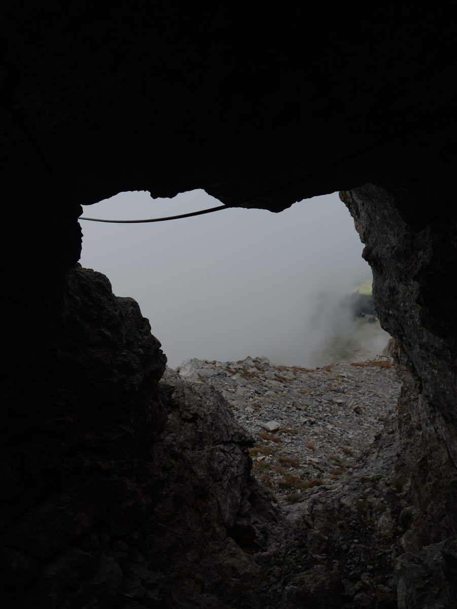Blick aus einer dunklen Felshöhle hinaus in den Nebel, ein Drahtseil spannt sich über die Öffnung