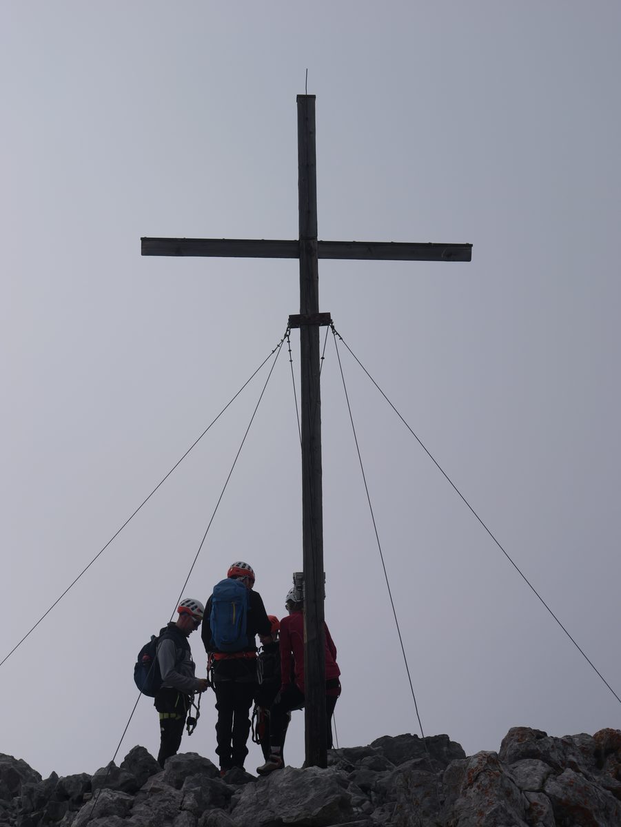 Hohes Gipfelkreuz als Silhouette vor grauem Himmel, darunter eine kleine Gruppe Bergsteiger