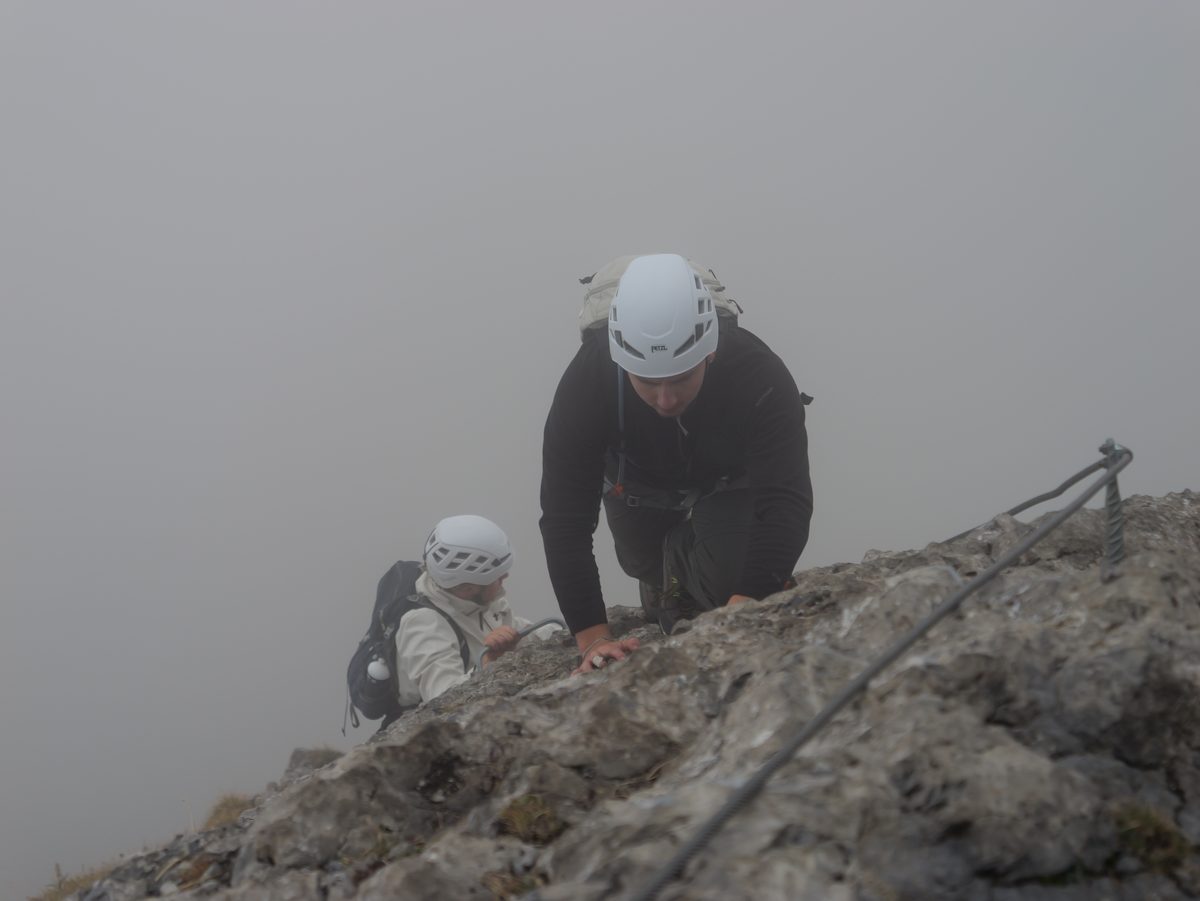 Zwei Kletterer mit weissen Helmen steigen am Seil über einen Felsrücken im dichten Nebel